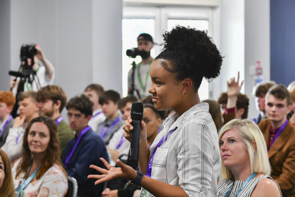 A student stands up to ask a question. She is holding a microphone and the crowd around her are listening intently