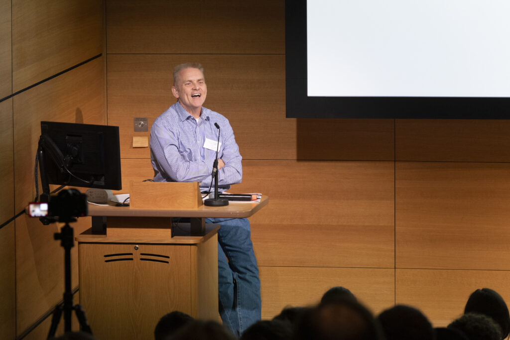 Professor Tom Welton OBE giving a presentation on stage to an audience. Smiling, and sitting down at the presenters desk.
