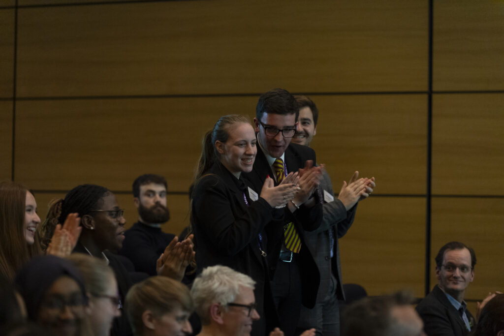 Three students stood clapping and celebrating surrounded by other students and teachers applauding in an awards ceremony.