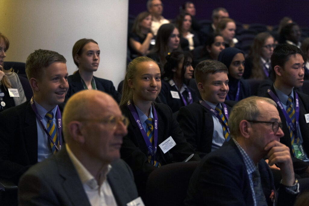 Students, teachers and staff watching a presentation, with one student smiling and facing the camera