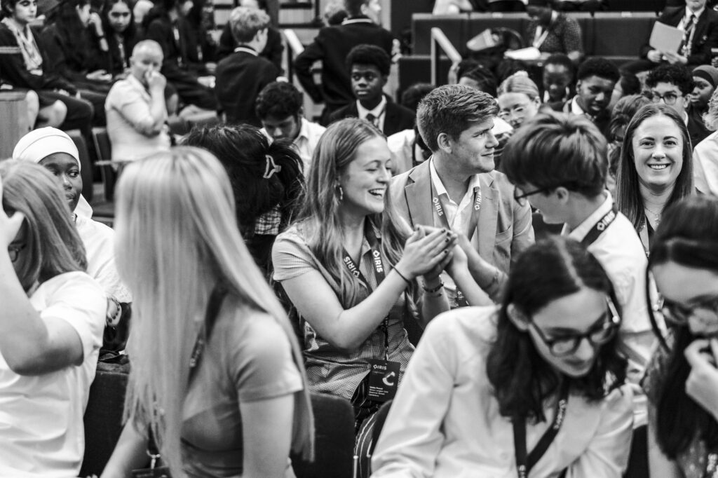 Students at an IRIS conference. They are seated in a hall talking to each other. Most of them are smiling and animated, some are looking at the camera.
